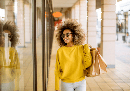 Mulher negra de óculos escuro, calça branca e blusa amarela, carregando sacolas de compra e andando na frente de lojas.