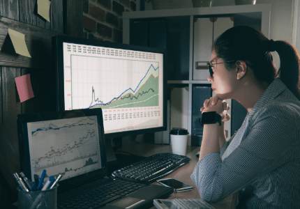 Mulher analista observando gráficos em duas telas de computador.