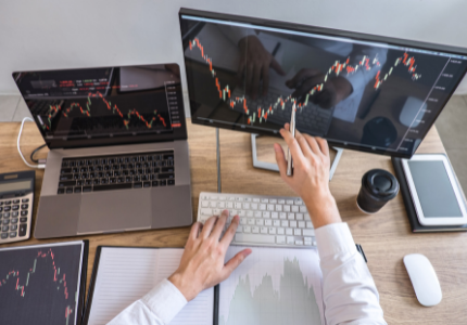 Pessoa sentada à mesa com material de trabalho, apontando para gráficos da Bolsa de Valores em seu computador.