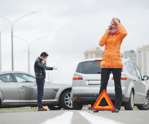 Dois carros que bateram e mulher ligando para o seguro, atrás de um triângulo.