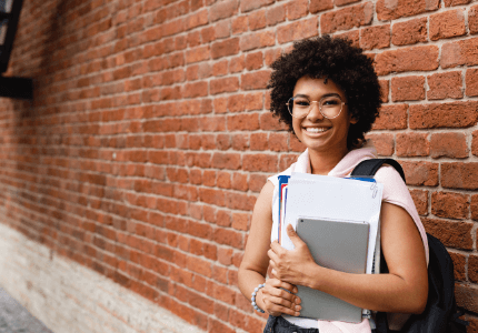 Estudante, negra e de óculos, feliz com seus livros no braço e mochila nas costas em frente a um muro de tijolo.