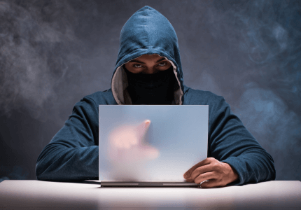 Hacker com capuz e máscara usando laptop para aplicar golpes em vítimas que querem contratar um empréstimo.
