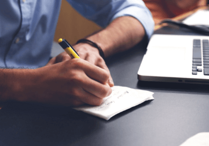 Homem de camisa social escrevendo em papel sobre mesa preta e notebook ao lado.