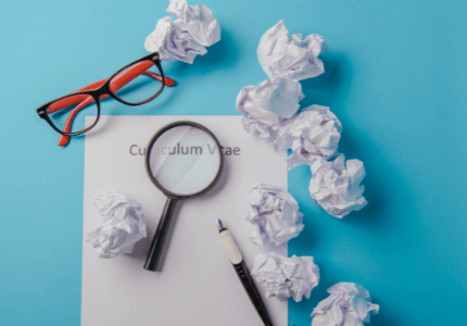 Óculos vermelho e preto em cima de uma mesa onde tem uma folha escrito Curriculum Vitae e uma lupa sobre a frase. Em volta, várias folhas de papel amaçadas.