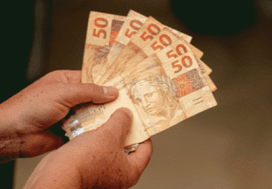 Duas mãos segurando seis notas de cinquenta reais.