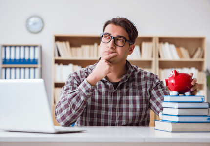 Homem sentado em frente a uma mesa, com a mão no queixo, pensativo sobre as taxas de juros. Em cima da mesa uma pilha de livros e em cima um cofre vermelho em forma de porquinho. Um laptop do outro lado da mesa.