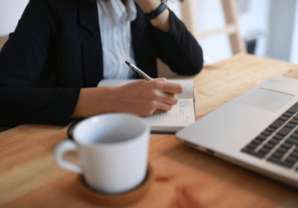 Pessoa sentada em uma mesa que tem uma caneca e um laptop. Ela está anotando em um caderno pequeno as 5 diferenças entre o saque imediato e o saque aniversário do FGTS.