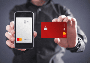 Mãos de um homem segurando um celular com um cartão de crédito do C6 Bank na cor prata e na outra mão um cartão de crédito C6 Bank na cor vermelha. Imagem representando uma análise completa ao cartão.