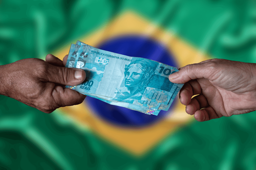 Mão entregando e a outra recebendo o dinheiro referente ao Auxílio Brasil. Ao fundo a bandeira do Brasil.