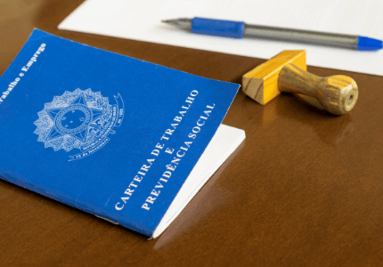 Carteira de trabalho brasileira, um carimbo e uma caneta em cima de um papel. Todos estão sobre uma mesa e representam a diminuição do desemprego no Brasil.