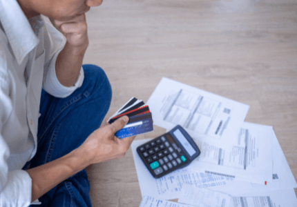Homem preocupado com as dívidas sentado ao chão, segurando cartões de crédito, olhando para boletos e uma calculadora
