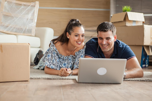 Casal de um homem e uma mulher, felizes, debruçados no chão, pedindo um empréstimo online pelo computador.