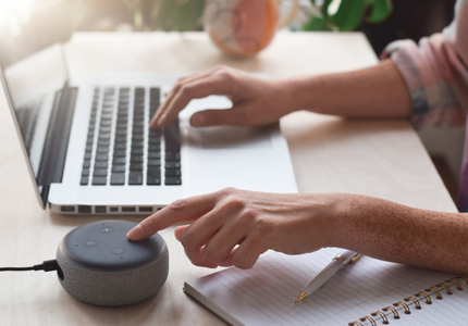 Braços de uma pessoa sobre a mesa, onde uma mão está no laptop e outra na Alexa da Amazon.