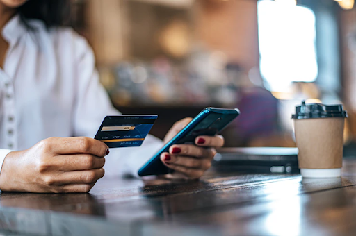 Mãos de uma mulher, apoiadas sobre a mesa segurando um cartão de crédito e um celular.