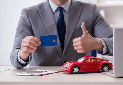 Homem de terno sentando em uma mesa, fazendo sinal de positivo com a mão esquerda e com a mão direita ele está segurando o cartão de crédito com que ele vai parcelar o pagamento do IPVA. Sobre a mesa um carrinho de miniatura representando o seu veículo.