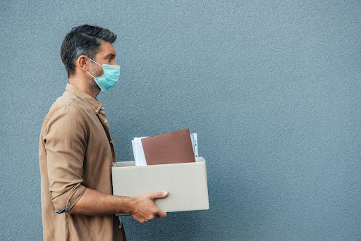 Homem com máscara cirúrgica, saindo do emprego que foi demito, segurando uma caixa com seus materiais.
