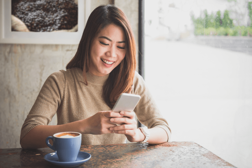 Mulher sorridente mexendo num celular, ao lado de uma xícara de café.
