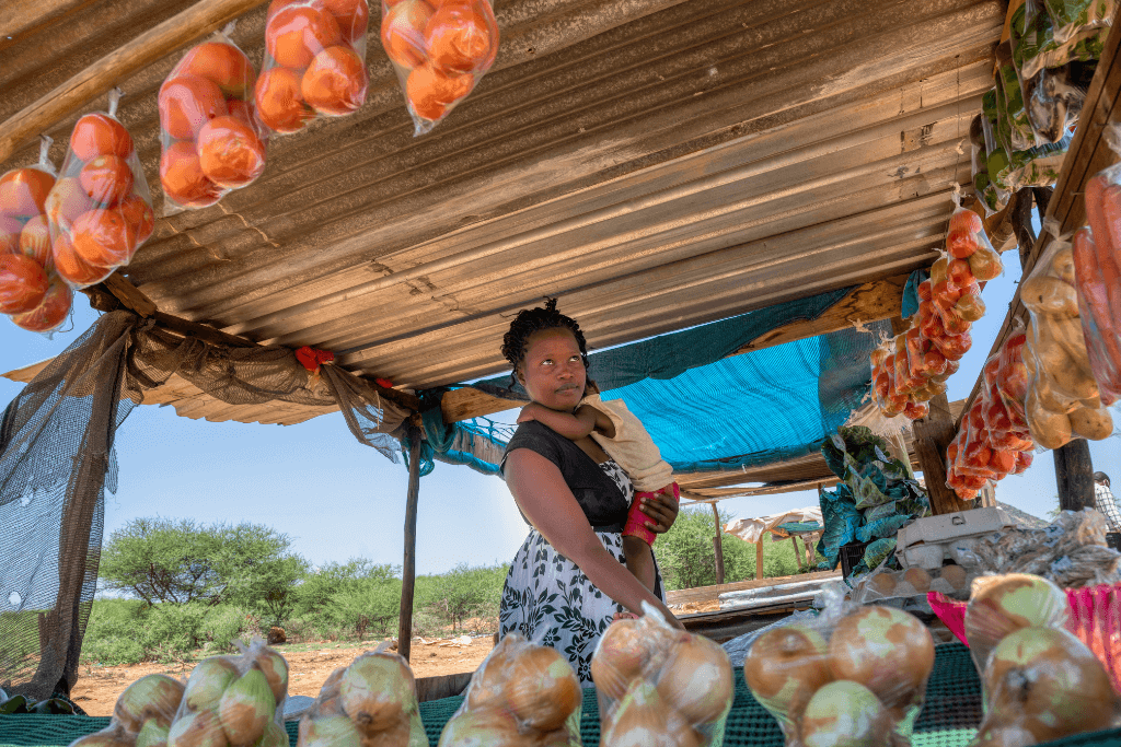 Mulher com criança no colo, vendendo produtos naturais numa barraca improvisada.