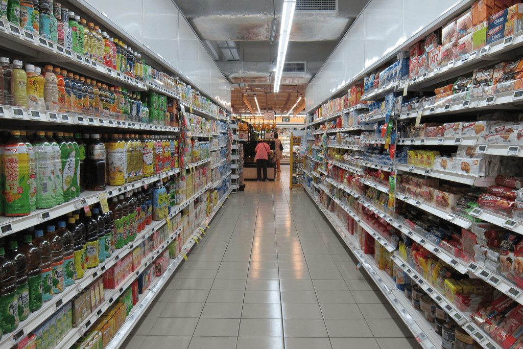 Supermercado cheiro de produtos, com mulher empurrando um carrinho ao fundo do corredor.