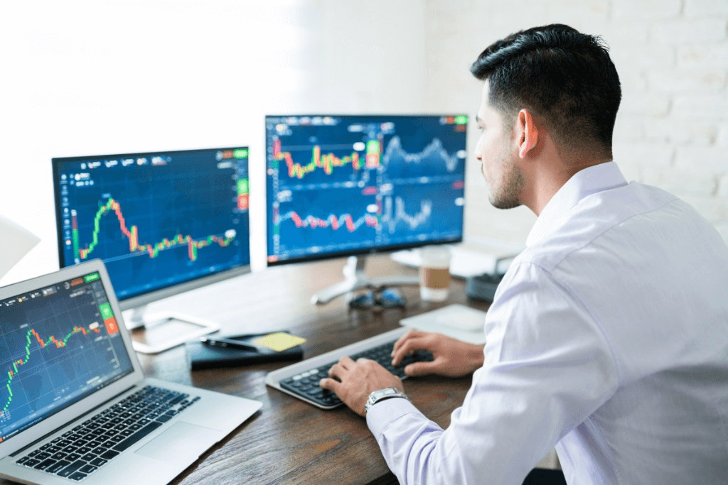 Homem acompanhando gráfico da bolsa de valores em três computadores diferentes.