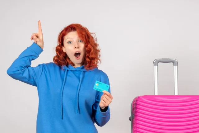 Mulher turista segurando cartão com programa de milhas Livelo ao lado de mala de viagem.